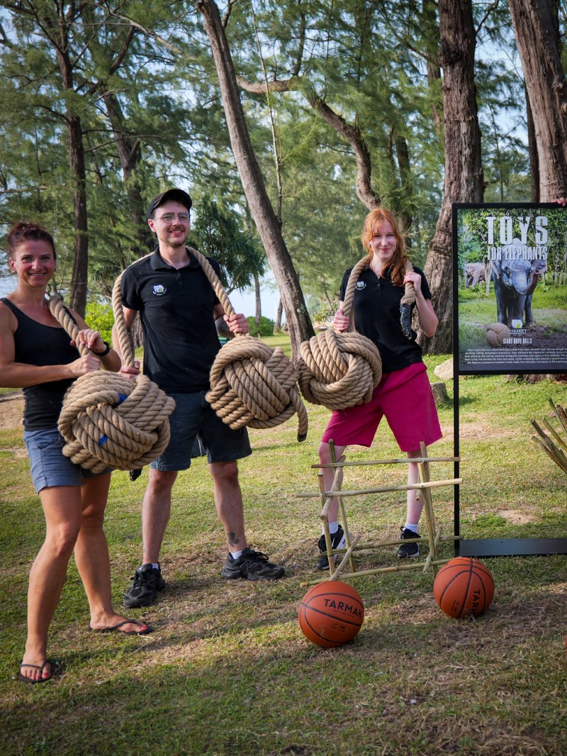 Three people outdoors holding large ropes near a display and basketballs on the ground.